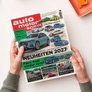 Von zwei Händen gehaltenes Magazin „auto motor sport“ mit rotem Titelbereich und weißem Schriftzug. Das Cover zeigt mehrere Autos sowie die Texte „NEUHEITEN 2027“, „ELFER GEGEN CORVETTE“, „Fährt Audi wieder vorn?“, „ALPENÜBERQUERUNG“ und „MERCEDES-S-KLASSE“; oben rechts liegt ein orangefarbenes Bürogerät.