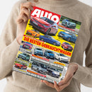 Eine Person mit beigem Strickpullover hält eine schräg ausgerichtete, glänzende Autozeitschrift mit rotem, gelbem und schwarzem Cover vor hellem Hintergrund. Sichtbar sind zahlreiche Fahrzeugfotos sowie Texte wie „AUTO“, „JETZT SPAREN BEIM AUTOKAUF! DIE BESTEN GEBRAUCHTWAGEN“, „Stärkster Serien-Porsche aller Zeiten“ und „Zwei kleine Raumwunder jagen den VW Polo“.