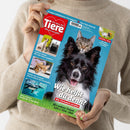 Person in beigem Strickpullover hält das Tiermagazin „Ein Herz für Tiere“ mit rotem Herz-Logo oben links. Titelthema: „Wie heißt du denn? Was Tiernamen verraten“, darunter ein schwarz-weißer Hund mit herausgestreckter Zunge und eine graue Katze dahinter, vor blauem Hintergrund. Linke Spalte mit kleineren Bildern von Katze, Hundebox, Meeresschildkröte und Texten wie „Endlich Urlaub! Mit oder ohne Haustier?“ und „Meister der Tarnung: Warum Katzen sich verstecken“.