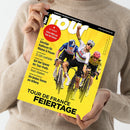 Person in beigefarbenem Pullover hält ein Magazin mit gelbem Titelblatt. Auf dem Cover sind drei männliche Radrennfahrer in Wettkampfausrüstung auf Rennrädern zu sehen, einer hebt den Arm zum Jubel. Schwarzer Schriftzug „Tour“ oben, rote runde Markierung mit „64 Seiten! Extra-Heft zur Tour de France“, Haupttitel „Tour de France Feiertage“, weitere Texte: „Günstige Radhosen für Männer & Frauen“, „Team Telekom“, „32-Zoll-Laufräder“ und „Die besten Touren am Arlberg“.