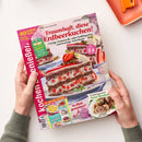 Titelblatt der Zeitschrift „Kochen & Genießen“, gehalten von zwei Händen über hellem Tisch. Hauptmotiv ist ein Stück Schichttorte mit Erdbeeren, Sahne und Schokoladenböden auf Teller, begleitet vom Schriftzug „Traumhaft, diese Erdbeerkuchen!“. Weitere sichtbare Texte: „Schwarzwälder Erdbeertorte“, „Frühlingsleicht Neue Rouladen“, „16 Seiten extra!“ und „So flink gezaubert!“. Hintergrund zeigt verschwommen weitere Kuchenscheiben.