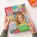 Titelbild der Zeitschrift „Bild Woche“ mit großem roten Logo oben links. In der Mitte eine lächelnde hellhaarige Frau vor hellblauem Hintergrund. Umgeben von bunten Textfeldern mit Schlagzeilen zu Themen wie „Die leckere Schlank-Lasagne“, „Wir lieben Käsekuchen!“, „Ein gesundes Herz in 8 Schritten“ und „Sicher unterwegs mit dem E-Bike“. Zwei Hände halten das Heft vor einem hellen Tisch mit orangefarbenem Gegenstand rechts oben.