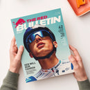 Titelseite des Magazins „The Red Bulletin“ mit großem Porträt eines männlichen Radfahrers in weiß-blauem Trikot, mit Helm und verspiegelter Sonnenbrille vor türkisfarbenem Hintergrund. Sichtbare Texte: „THE RED BULLETIN“, „DER WILL NUR FAHREN“, „42 Seiten Sommer-Special: Feel the Real“, Logos u. a. von Red Bull, Bora und UCI World Team. Zwei Hände halten das Magazin über einer weißen Fläche mit orangener Schale.