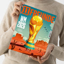 Person in beigem Strickpullover hält eine Ausgabe des Magazins „11 FREUNDE“ mit türkis-orangem Titelblatt; mittig eine Illustration des WM-Pokals in gold auf Wüstenlandschaft mit Felsen und Kakteen, darüber Text „WM 2026“ sowie Unterzeile „Das Sonderheft zur Weltmeisterschaft in Mexiko, Kanada und den USA“, im Hintergrund drei fliegende Jets und leichter Wolkenhimmel.