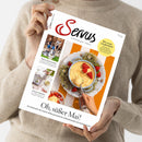 Zwei Hände einer Person in beigefarbenem Strickpullover halten eine Ausgabe der Zeitschrift „Servus – einfach gut leben“. Auf dem Titelbild: eine Schale mit gelbem Eis und frischen Erdbeeren auf orange-weißem Untergrund, dazu kleinere Fotos von Menschen, einem gedeckten Tisch und Gebäck. Sichtbare Texte u. a.: „Oh, süßer Mai!“, „Wie werde ich glücklich?“ sowie Themenhinweise zu Wohnen, Heimat und Rezeptideen.