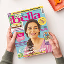 Aufgeschlagene Zeitschrift „bella“ auf weißer Fläche, von zwei Händen mit grauen Strickärmeln gehalten; auf dem Cover eine lächelnde Frau mit braunem Haar vor türkisem Hintergrund. Sichtbare Texte: „KOCHEN Raffinierte Blitz-Rezepte“, „Das tut mir gut“, „Nur Mut! Schnell zu mehr innerer Kraft“, „Cremige Eierlikör-Torte“ und „Die neuen Super-Suppen“. Oben rechts liegt ein orangefarbenes Bürogerät.