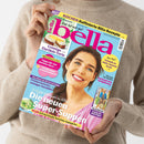 Weibliche Person im beigefarbenen Strickpullover hält eine schräg zur Kamera ausgerichtete Zeitschrift vor hellem Hintergrund; das farbige Cover zeigt eine lächelnde Frau, ein Kuchenfoto und weitere kleine Bilder. Lesbare Aufschriften: „bella“, „Kochen: Raffinierte Blitz-Rezepte“, „Das tut mir gut“, „Cremige Eierlikör-Torte“, „Medizin-Extra: Tipps für ein starkes Herz“ und „Die neuen Super-Suppen“.