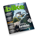 Schräg fotografiertes, glänzendes „bike“-Magazin auf schwarzem Hintergrund mit großem grünem Titel „bike“ und Aufschrift „DEINE NR.1“. Das Cover zeigt eine männliche Person auf einem gelben Mountainbike beim Sprung über felsiges Gelände; lesbare Überschriften sind „Gabel-Duell“, „Canyon gegen YT und Radon“, „Kids-Stuff“, „Bikepacking“, „Eurobike“ und „TARIFA“.