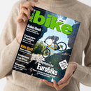 Eine Person ohne sichtbares Gesicht, deren Geschlecht nicht eindeutig erkennbar ist, hält ein schräg ausgerichtetes Magazin im grünen Strickpullover. Das Cover zeigt einen Mountainbiker auf gelbem Fahrrad vor einer Felslandschaft und trägt die Aufschrift „bike“ sowie Texte zu Gabel-Duell, Trailbikes, Bikepacking, Kids-Stuff und den Highlights der Eurobike.