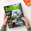Aufgeschlagenes Mountainbike-Magazin „bike“ mit großem grünen Titel, gehalten von zwei Händen über weißer Fläche. Titelbild mit einer Person auf gelbem Mountainbike vor Felslandschaft; lesbare Texte: „DEINE NR. 1“, „Gabel-Duell“, „Kids-Stuff“, „Bikepacking“, „Die Highlights der Eurobike“ und „Tarifa – Die besten Trails an Spaniens Südküste“.