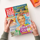 Titelseite der Frauenzeitschrift „Bild der Frau Woche“, von zwei Händen gehalten. Das Cover zeigt eine blonde weibliche Person, die Überschrift „TRUE CRIME“, den Hinweis „Tatort Promi-Welt“, ein TV-Programm, einen Käsekuchen, Blumensträuße sowie weitere Schlagzeilen zu Ernährung und Schlank-Eis; oben rechts liegt ein orangefarbenes Objekt auf weißem Untergrund.