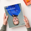 Auf weißer Fläche halten zwei Hände mit grauen Strickärmeln ein schräg liegendes Magazin. Das Cover zeigt eine weibliche Person mit blonden Haaren und blauen Augen in leuchtend blauer Kleidung; lesbare Texte sind „brand eins“, „Das geht!“, „Wie du mich siehst, bestimme ich.“, „brandeins.de“ und „Schwerpunkt Schönheit“. Oben rechts liegt teilweise ein orangefarbenes Objekt.