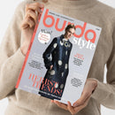 Weibliche Person im beigefarbenen Strickpullover hält eine schräg fotografierte Zeitschrift mit hellgrauem Cover und korallenroter Typografie. Lesbare Texte: „burda style“, „NÄHEN AUS LEIDENSCHAFT“, „HERBST TRENDS“, „ICONIC FASHION MOMENT“, „DEEP PURPLE“, „POWER LOOKS“ und „Die schönsten Schnitte der Saison zum Nachnähen“; abgebildet ist eine Frau im dunkelblauen Mantel mit weißen Kreisen.