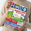 Eine Person in beigefarbenem Strickpullover hält das Technikmagazin „PC-WELT+“ Ausgabe 8/2026 vor neutralem Hintergrund; das Cover zeigt Schlagzeilen wie „Registry-Tricks mit Sofortwirkung“, „Windows nach Maß“, „Anti-Phishing-DVD“, „Windows 11 Pannenhilfe“ und mehrere farbige Textfelder in Rot, Gelb, Blau und Grün mit Abbildungen von Computerbildschirmen, DVDs und Software-Symbolen.