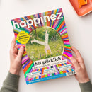 Titelblatt des Mindstyle-Magazins „happinez“ mit bunter, kreisförmig strahlenförmiger Hintergrundgestaltung in Regenbogenfarben. In der Mitte eine Illustration einer Person, die im Gras einen Handstand macht, umgeben von Blumen. Sichtbare Texte: „Sei glücklich – Alles strahlt heller, wenn Du lachst“, gelbe Plakette mit „EXTRA Zauberhafte Lesezeichen für die Urlaubslektüre“ sowie Themenfelder zu Selbst-Erkenntnis, Liebes-Magie, Aktiver Hoffnung und Weiblichkeit.