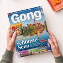 Magazintitel „Gong“ mit großem blauem Schriftzug auf hellblauem Hintergrund, darüber Datumsangabe vom 24.5. bis 30.5.2025. Titelthema: „Deutschlands schönste Seen“, mit Unterzeile „Plus Österreich“. Im Hintergrund Schloss mit Türmen und goldenen Reflexionen im Wasser. Zusätzliche Texte: „Raffinierte Rezepte mit Hack“, „Die Schule der kleinen Teddys“ und „Die neuen Tricks der Betrüger“. Zwei Hände halten das Magazin auf hellem Tisch.