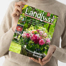 Person in beigem Strickpullover hält ein Magazin mit grünem Titelbild „Landlust“ in den Händen. Auf dem Cover ein großer Strauß aus rosa, roten und weißen Rosen mit grünen Blättern, im Hintergrund unscharfe Gartenlandschaft. Sichtbare Texte: „20 Jahre Landlust“, „Staudenbeete anlegen und gestalten“, „Eifel: Auf zu Ginsterblüte und Vulkanen!“, „Basteln mit Papier und Salzteig“, unten „Rosenfreude“.