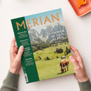 Magazin „MERIAN – The Art of Travel“ mit Titelthema „Sommer in den Alpen“, abgebildet ist eine grüne Berglandschaft mit grasender Kuh und Hütten vor Felsen im Hintergrund; linke Spalte mit Texten zu Reisezielen „Mexico City“, „Korfu & Co“, „München“ und „Teneriffa“; gehalten von zwei Händen über hellem Hintergrund mit orangefarbenem Gegenstand am oberen Bildrand.
