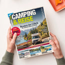 Schräg fotografierte Zeitschrift „CAMPING & REISE“ mit dem Titel „Routen ins Glück – Die schönsten Reisen in Europa“, gehalten von zwei Händen vor weißem Hintergrund. Das Cover zeigt ein Wohnmobil vor einer mediterranen Berglandschaft sowie Hinweise auf „Caravan Salon 2026“, „Hartschalen-Dachzelt“, „Vogtland“, „Münsterland“, „Südtirol“, „Auvergne“ und „Finnland“; oben rechts liegt ein orangefarbenes Objekt.