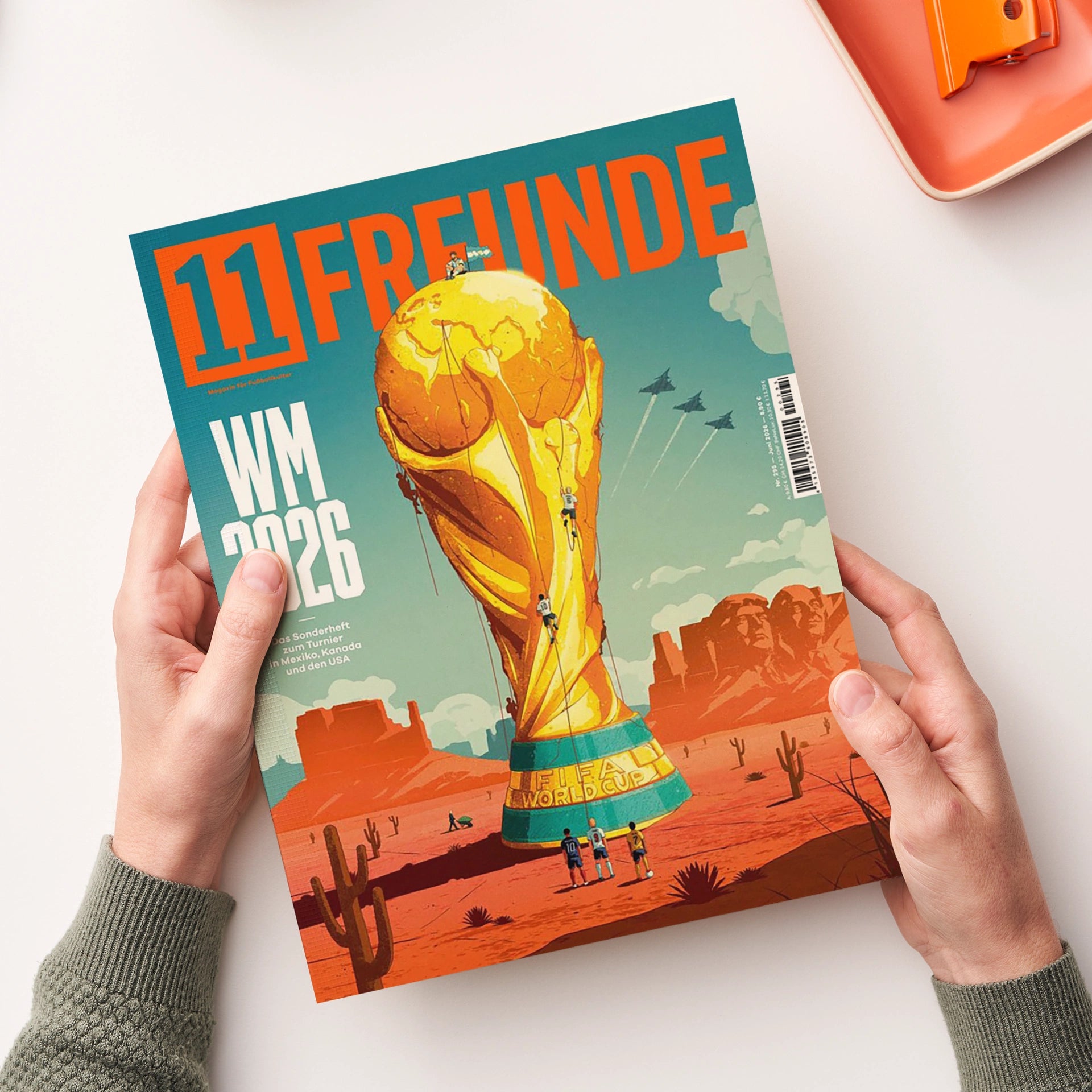 Magazintitel „11 FREUNDE“ in großer oranger Schrift auf türkisfarbenem Hintergrund, mit der Aufschrift „WM 2026“ links unten. Illustration einer riesigen goldenen WM‑Trophäe in einer Wüstenlandschaft mit Kakteen, kleinen Figuren und Flugzeugen am Himmel, im Hintergrund Bergformationen und das Mount‑Rushmore‑Relief; gehalten von zwei hellhäutigen Händen vor hellem Tischhintergrund.
