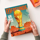 Magazintitel „11 FREUNDE“ in großer oranger Schrift auf türkisfarbenem Hintergrund, mit der Aufschrift „WM 2026“ links unten. Illustration einer riesigen goldenen WM‑Trophäe in einer Wüstenlandschaft mit Kakteen, kleinen Figuren und Flugzeugen am Himmel, im Hintergrund Bergformationen und das Mount‑Rushmore‑Relief; gehalten von zwei hellhäutigen Händen vor hellem Tischhintergrund.