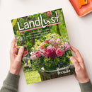 Titelseite der Zeitschrift „Landlust“ mit großem Blumenarrangement aus rosafarbenen, roten und weißen Rosen in grünem Eimer auf Holztisch, umgeben von grüner Wiese. Aufschrift u. a. „Rosenfreude“, „Staudenbeete anlegen und gestalten“, „Eifel: Auf zu Ginsterblüte und Vulkanen!“, „Basteln mit Papier und Salzteig“, Logo „20 Jahre Landlust“ oben rechts. Gehalten von zwei Händen über hellem Hintergrund.