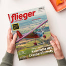 Titelblatt des „flieger MAGAZIN“ in roter Schrift auf hellblauem Hintergrund mit Luftaufnahme eines roten einmotorigen Flugzeugs über ländlicher Landschaft. Sichtbare Textzeilen: „BLEIFREIES AVGAS – Die erste Tankstelle in Ganderkesee“, „Oldtimer-Porträt: Cessna 140 – Keimzelle der Cessna-Einmots“, „AERO 2025 – Alle News & Trends der Messe“, „BALTIKUM-REISE – Unberührte Natur und einsame Inseln“, „PRAXIS-TIPPS – Sicher fliegen im Gebirge“. Zwei Hände halten die Zeitschrift.