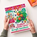 Titelbild des Magazins „Good Health“ mit farbenfroher Gestaltung, großer Überschrift „Endlich wieder neue Kraft tanken“ und Bildarrangement aus Spinatblättern, Tomaten, Blaubeeren, Walnüssen und Mandeln. Oben links ein rundes Foto zweier weiblich gelesener Personen beim Radfahren mit dem Text „Wie Bewegung unser Gehirn fit hält“. Im unteren Bereich eine kleine Abbildung eines Joghurt-Glases mit Kirschen vor hellem Hintergrund.