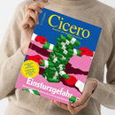 Person in beigefarbenem Strickpullover hält ein schräg abgebildetes Magazin mit blau-pinkem Cover. Der Titel „Cicero“ und der Untertitel „Magazin für politische Kultur“ sind sichtbar; das Cover zeigt rote Handschuhe, gestapelte grüne Geldscheine sowie die Texte „Sehnsucht oder Albtraum? Großbritannien streitet über die Abkehr vom Brexit“ und „Einsturzgefahr“.