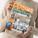Eine weibliche Person in beigefarbenem Strickpullover hält schräg ein Magazin mit orangefarbenem „COUCH“-Titel. Das Cover zeigt „FERIENFLAIR ZU HAUSE“, eine Frau mit kleinem Hund in einem mediterran eingerichteten Zimmer sowie die Texte „HOMESTORYS“, „Coolcation“, „Strandgut upgraden“ und „Perfekte Reisebegleiter“.