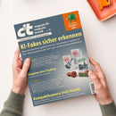 Schräg gehaltenes, blau-graues c’t-Magazin mit dem Text „magazin für computer technik“ und „Europas größtes IT- und Tech-Magazin“. Sichtbare Titel: „KI-Fakes sicher erkennen“, „Shopping ohne Tracking“ und „Kompaktkamera trotz Handy“; daneben Illustrationen zu KI-Kennzeichnungen. Zwei Hände mit grünen Ärmeln halten das Heft, das Geschlecht der Person ist nicht erkennbar. Oben rechts liegen ein oranges Tablett und ein orangefarbenes Gerät.