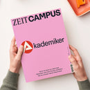 Titelblatt der Zeitschrift „ZEIT CAMPUS“ mit hellrosa Hintergrund, großem schwarzen Schriftzug „Akademiker“ und rotem Kreis mit weißem Dreieck-Symbol links daneben. Unten steht der Text: „Noch nie waren so viele junge Menschen nach dem Studium arbeitslos. Wie du trotzdem deinen Weg findest.“ Zwei Hände in graugrünem Pullover halten das Magazin über hellem Untergrund, oben rechts liegt eine orangefarbene Schale.