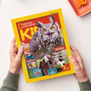 Titelseite des Magazins „National Geographic Kids“ mit gelbem Rand und rotem Hintergrund, im Mittelpunkt eine Eule mit geöffnetem Schnabel. Text: „EULEN: Mit großen Augen durch die Nacht!“, „2 starke Poster!“, „Orcas – ihr geheimes Leben!“, „Pharaonen – Herrscher über das alte Ägypten!“, „Dinosaurier – Unglaubliches über die Urzeittiere!“. Gehalten von zwei Händen über heller Tischfläche.