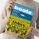 Person in beigem Strickpullover hält das Magazin „boote“, Ausgabe 5/2025, vor neutralem Hintergrund. Titelbild zeigt ein gelbes Rapsfeld im Vordergrund, dahinter die blaue Ostsee mit einem weißen Motorboot und mehreren kleinen Segelbooten. Aufschrift: „Revier-Update Ostsee – Deutschland, Polen und Dänemark“, weitere Texte zu Themen wie Gebrauchtboote, Wakeboard, Törnplanung und Führerschein.