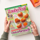 Titelblatt des Magazins „Zaubertopf – Lieblingsrezepte für Thermomix“ in grüner und violetter Farbgestaltung, gehalten von zwei Händen über hellbeigem Tisch. Abgebildet sind fünf herzförmige Hefegebäcke mit Marmelade und Butter auf grünem Teller. Textelemente u. a.: „Liebevolle Ideen: Osterbrunch“, „Frühlingsfit! Alles strahlt!“, „Eierlikör“, „Hollandaise“, „Hefe-Herzen mit Marillenfruchtaufstrich“.
