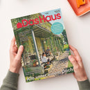 Zeitschrift „Das Haus“ mit rotem Titel auf hellblauem Hintergrund, gehalten von zwei Händen. Das Cover zeigt ein bepflanztes Gewächshaus mit einer erwachsenen Person im gestreiften Shirt, einer weiteren erwachsenen Person und einem Kind; Texte: „Bauen Wohnen Schöner Leben“, „Naturgarten: So schaffen Sie Lebensräume für Wildbienen, Igel und Co.“, „Farben & Tapeten“, „Solartechnik“ und „Alle unter einem Dach“.