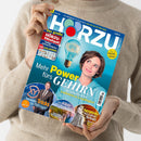 Person in beigefarbenem Strickpullover hält die Zeitschrift „HÖRZU“ mit blau-türkisem Cover. Zentral ein Porträt einer braunhaarigen Frau neben einer leuchtenden Glühbirne, Überschrift: „Mehr Power fürs Gehirn – So verbessern Sie Gedächtnis & Lernfähigkeit“. Weitere Texte: „Die neuen Tricks der Betrüger“, „Ägyptens wertvollster Schatz“, „Eine Kiste zum Verlieben“, gelbes Siegel „Nr. 1 Deutschlands erstes TV-Magazin“.