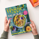 Titelblatt der Zeitschrift „slowly veggie! – vegetarisch & vegan genießen“ mit dem Haupttitel „Rund um die Welt“. Mittelpunkt des Covers ist eine Schale mit Gemüse-Couscous, Zucchini, Karotten, Nüssen und essbarer Blüte, daneben Essstäbchen. Weitere sichtbare Texte: „Super Greens“, „Urban Jungle“, „Rezepte, die glücklich machen“. Gehalten von zwei Händen vor hellem Hintergrund mit einem orangefarbenen rechteckigen Gegenstand oben rechts.