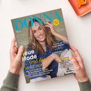 Schräg von oben fotografierte „DONNA“-Zeitschrift, von zwei Händen über einer weißen Fläche gehalten. Auf dem Cover: lächelnde Frau mit braunem Haar und Jeanshemd, gelber Kreis „Meine Zeit ist jetzt“ sowie Texte wie „Lesen, lachen, abtauchen“, „neue Mode“, „Zurück zur Jugendliebe?“ und „Andalusien“. Oben rechts liegt ein orangefarbenes Notizbuch mit Büroklammer.
