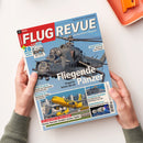 Titelbild der Zeitschrift „FLUG REVUE“ mit großem Foto eines grauen Militärhubschraubers Mi-24 im Flug vor blauem Himmel, Hauptüberschrift „Fliegende Panzer – Ungarns letzte Mi-24“. Weitere sichtbare Texte: „Zukunftsflügel mit KI und ohne Klappen“, „US-Airlines in der Krise“, „28 Seiten extra“. Gehalten von zwei Händen vor hellem Hintergrund mit orangefarbenem Objekt rechts oben.