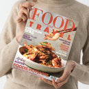 Eine Person in beigem Wollpullover hält das Magazin „Food and Travel“ in deutscher Ausgabe, mit Titel in roten Großbuchstaben. Auf dem Cover ein Teller mit gebratenen Garnelen in goldbrauner Kruste, darüber ein Garnelenstück in Essstäbchen. Sichtbare Textzeilen: „Spektakuläre Spas“, „Asiatische Küche“, „Rezepte von der Donau“, „Afrikas Küsten“ und „Foodie-Hotspots“.