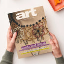 Titelblatt der Zeitschrift „art – Das Kunstmagazin / Juni 2023“, gehalten von zwei Händen über einer hellen Tischoberfläche; das Cover zeigt eine Nahaufnahme einer kunstvollen Schmuckgestaltung mit goldenen, schwarzen und roten Elementen, Perlen und Flechtmustern; sichtbare Überschriften u. a. „Glanz und Gloria“, „Edelsteine, Gold und Perlen: Eine kleine Kunstgeschichte des Schmucks“, „EXPO Osaka: Das Weltfestival der Nachhaltigkeit“.