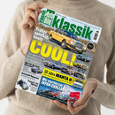 Eine Person in beigem Strickpullover hält die Zeitschrift „Auto Bild Klassik“ mit grün-weißem Logo und Titel „Früher spießig, heute COOL!“ in großen gelben Buchstaben. Auf dem Cover sind mehrere klassische Autos in unterschiedlichen Farben zu sehen, darunter Mercedes 230 W123, Opel Commodore C, Ford Granada 2.8, sowie Textfelder zu Themen wie „50 Jahre Manta B“ und „Japan-Challenge“.