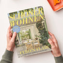 Titelblatt der Wohnzeitschrift „SCHÖNER WOHNEN“ mit gelber Schrift auf einem Foto eines hellen, natürlich eingerichteten Raumes mit Holztisch, weißen Stühlen, Pflanzen und geöffneten grünen Fensterläden; sichtbare Textaufdrucke wie „FERIEN-FLAIR“, „Sommerhäuser von Dänemark bis Marokko“, „Komfort-Zone“ und „Outdoor Tipps“; gehalten von zwei Händen mit grünem Strickpullover vor hellbeigem Hintergrund.