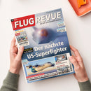 Titelblatt der Zeitschrift „FLUG REVUE“ Ausgabe 06/2025, gehalten von zwei Händen über hellorangefarbener Ablage; Haupttitel „Der nächste US-Superfighter“ mit unscharfem Kampfflugzeug im Nebel; kleinere Überschriften u. a. „Boeing F‑47: Erste Bilder“, „Technologie-Bausteine – Was plant Airbus für sein nächstes Flugzeug?“, „Ramstein Flag – Die große Übung der NATO“ und „Messe AERO 2025 – Business Jets am Bodensee“.
