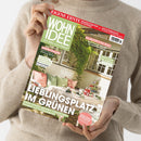 Helles Titelbild der Zeitschrift „Wohnidee“ mit grün-weißem Logo, gehalten von einer Person in beigem Pullover. Abgebildeter Terrassenbereich mit Tisch, Decke, Blumen, Gläsern und Kissen in Pastellfarben unter Rankpflanzen. Sichtbare Überschriften: „Lieblingsplatz im Grünen“, „Gesund schlafen“, „Wenig Platz viel Gefühl“, „Da blüht uns was!“ und „Versteckt, verschoben, verwandelt – Stauraummöbel neu gedacht“.