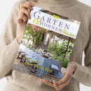 Heller Magazinumschlag der Ausgabe „Garten & Wohnen Träume 3/2025“, gehalten von einer Person in beigem Strickpullover vor neutralem Hintergrund. Titelmotive zeigen einen mediterranen Garten mit gedecktem Esstisch unter Sonnenschirm, blühende Sträucher und Sitzmöbel. Sichtbare Texte: „Ein Sommer zum Genießen“, dazu Themenhinweise zu mediterranen Aromen, Marktschätzen, Interieurs und Gärten der Toskana.