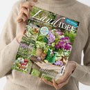 Frau in beigem Strickpullover hält die Zeitschrift „LandIDEE“ in beiden Händen, Titelthema „Bezaubernder Blütentraum im Sommer“. Auf dem Cover stehen ein grüner Metallkrug mit lila und weißen Fliederblüten auf einem Holztisch, daneben Tontöpfe und Gartenpflanzen. Weitere Aufschriften: „Leckere Sommerrezepte mit Erbsen“, „Erdbeeren & Co. für unsere Gesundheit“, „Kreative Ideen für den Balkon“, „Kugelförmige Blüten“.