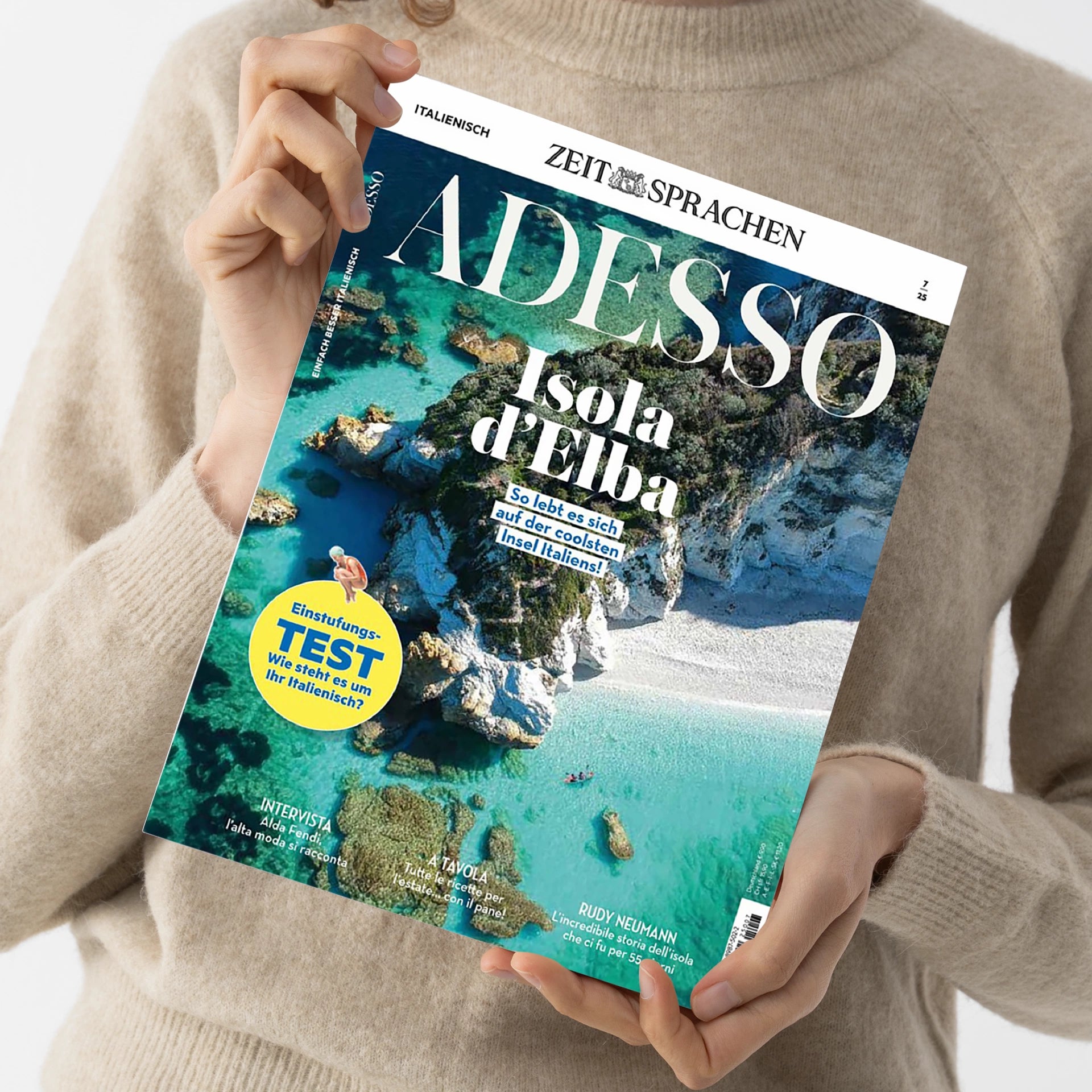 Eine Person in beigefarbenem Wollpullover hält eine Ausgabe des Magazins „Adesso“ mit dem Titelblatt „Isola d’Elba – So lebt es sich auf der coolsten Insel Italiens“. Sichtbar sind das Logo „ZEIT SPRACHEN“, ein gelber Kreis mit dem Text „Einstufungs-TEST – Wie steht es um Ihr Italienisch?“ sowie ein Luftbild der weißen Felsküste und des türkisblauen Meeres der Insel Elba.