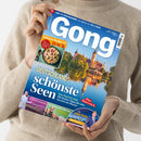 Helläufige Person in beigefarbenem Strickpullover hält die Zeitschrift „Gong“ mit blauem Titelblatt, großem weißen Schriftzug und dem Hauptthema „Deutschlands schönste Seen“. Oberer Bereich zeigt ein Foto eines Schlosses mit Spiegelung im Wasser, daneben runder Ausschnitt mit Gericht „Raffinierte Rezepte mit Hack“. Weitere Texte: „Wie gut ist Ihr Allgemeinwissen?“, „Die Schule der kleinen Teddys“, „Die neuen Tricks der Betrüger“.